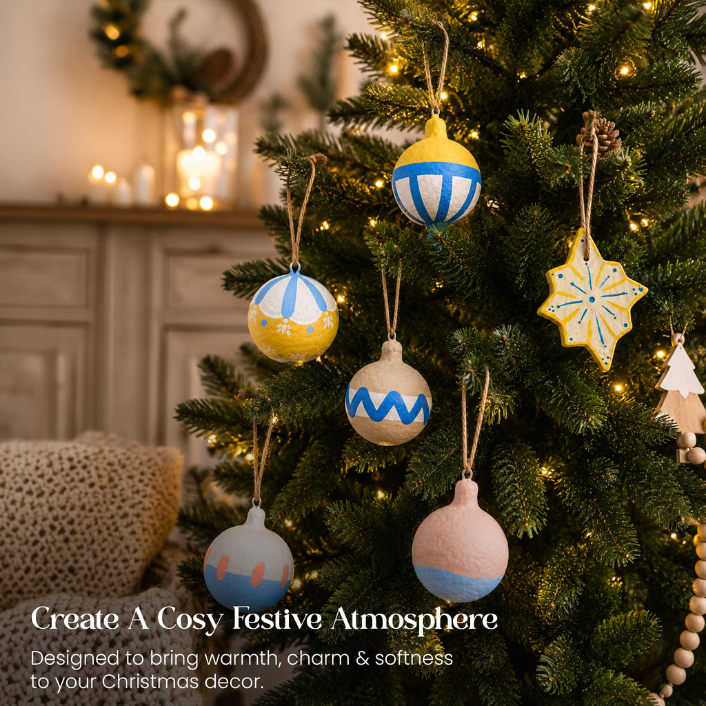 Artisan hand-painted paper mache Christmas ornaments hanging on tree in cosy living room with candles and fairy lights