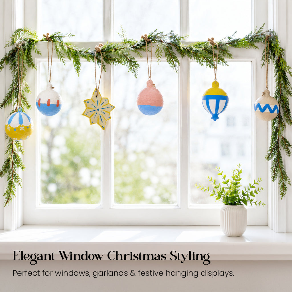 Handmade paper mache Christmas baubles hanging from pine garland on window — elegant Scandi festive window display UK