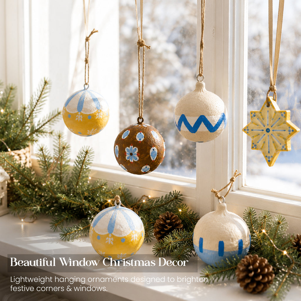 Hand-painted paper mache Christmas baubles hanging in snowy window display with pine garland and pine cones UK
