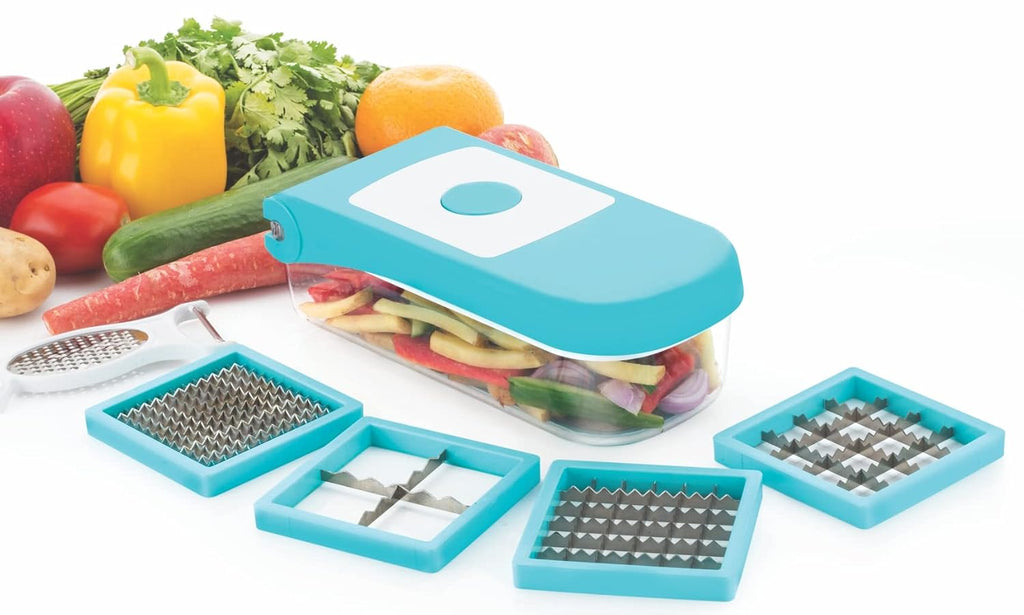 The dicer beside its four blade inserts and peeler-grater, its container filled with cut vegetables, fresh produce behind